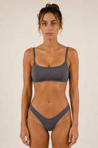 A woman modeling a two-piece Sulkii polewear set in a solid dark gray color, consisting of a scoop-neck bikini top and matching high-cut bikini bottoms, against a plain beige studio background.