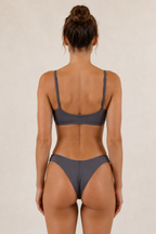 A woman modeling a two-piece Sulkii polewear set in a solid dark gray color, consisting of a scoop-neck bikini top and matching high-cut bikini bottoms, against a plain beige studio background.