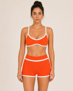 A woman modeling a bright orange two-piece sulkii polewear set with white trim, consisting of a sports bra and matching shorts, against a plain beige background.